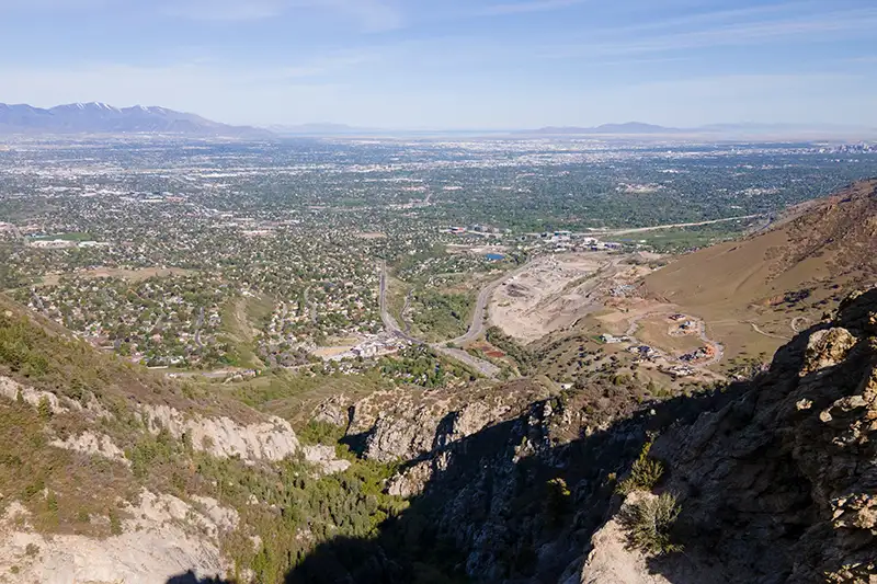 Sweeping view of a valley city and suburbs with distant mountains, seen from a rocky mountain ridge above a wooded canyon.