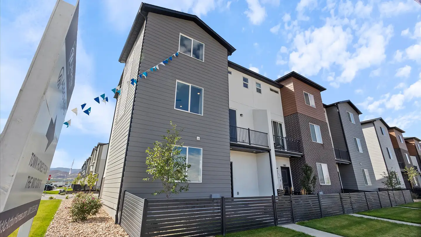 Three-story modern townhomes with dark gray siding, a brick accent, and an Arrowhead townhome sales sign with bunting flags.