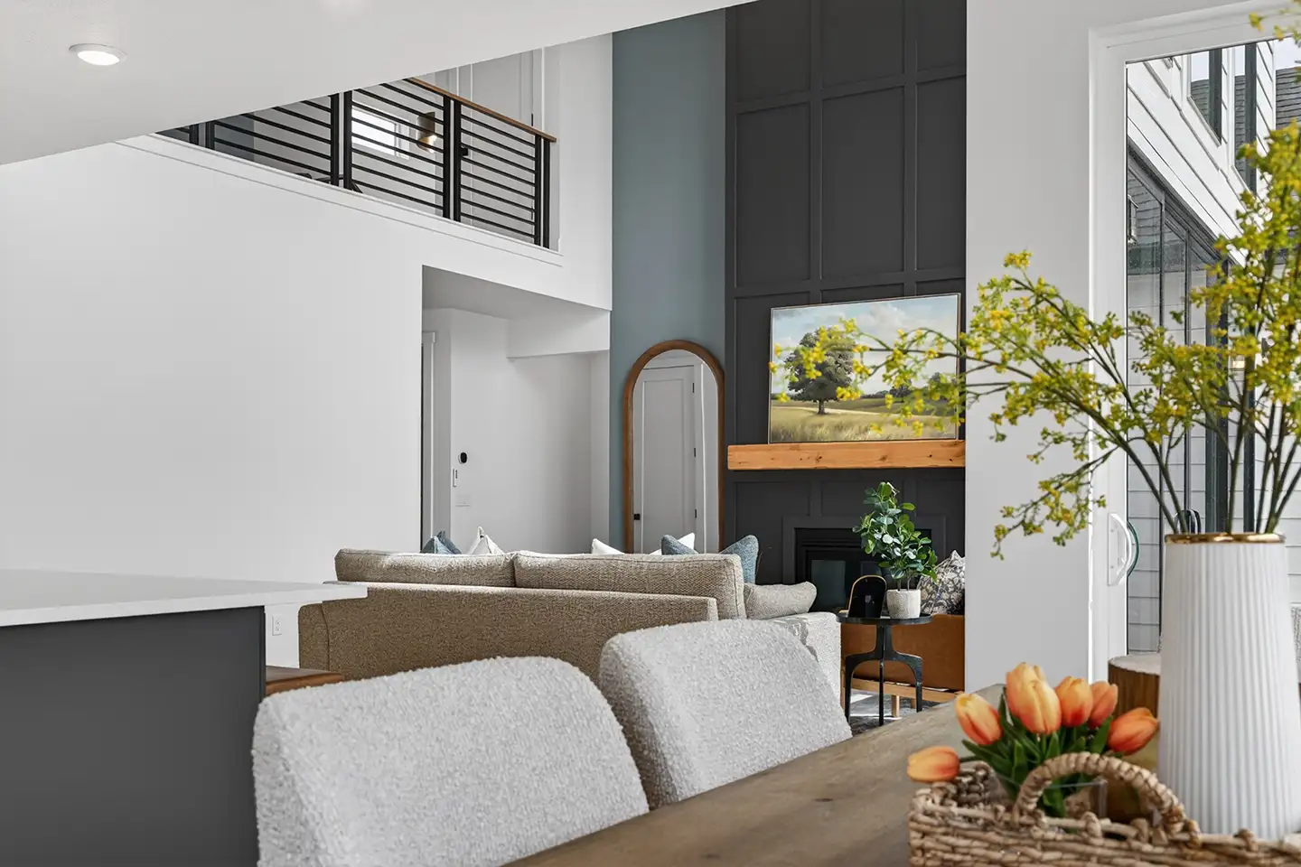 Two-story living room with vaulted ceiling, dark board-and-batten fireplace wall, framed landscape art, and neutral gray sofa seating.