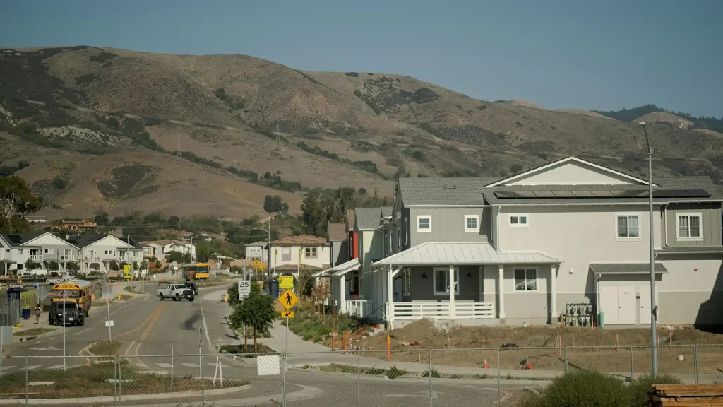New residential community under construction with finished homes, a school bus on the street, and dry foothills rising in the background.