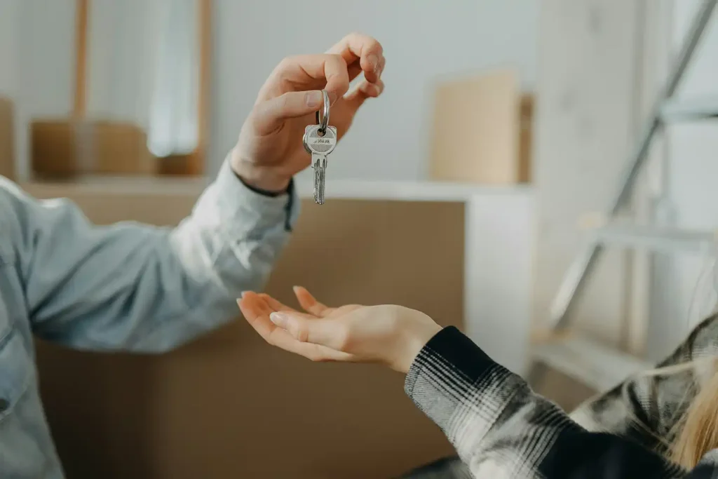 One person drops a set of house keys into another person's open palm, with moving boxes stacked in the background.