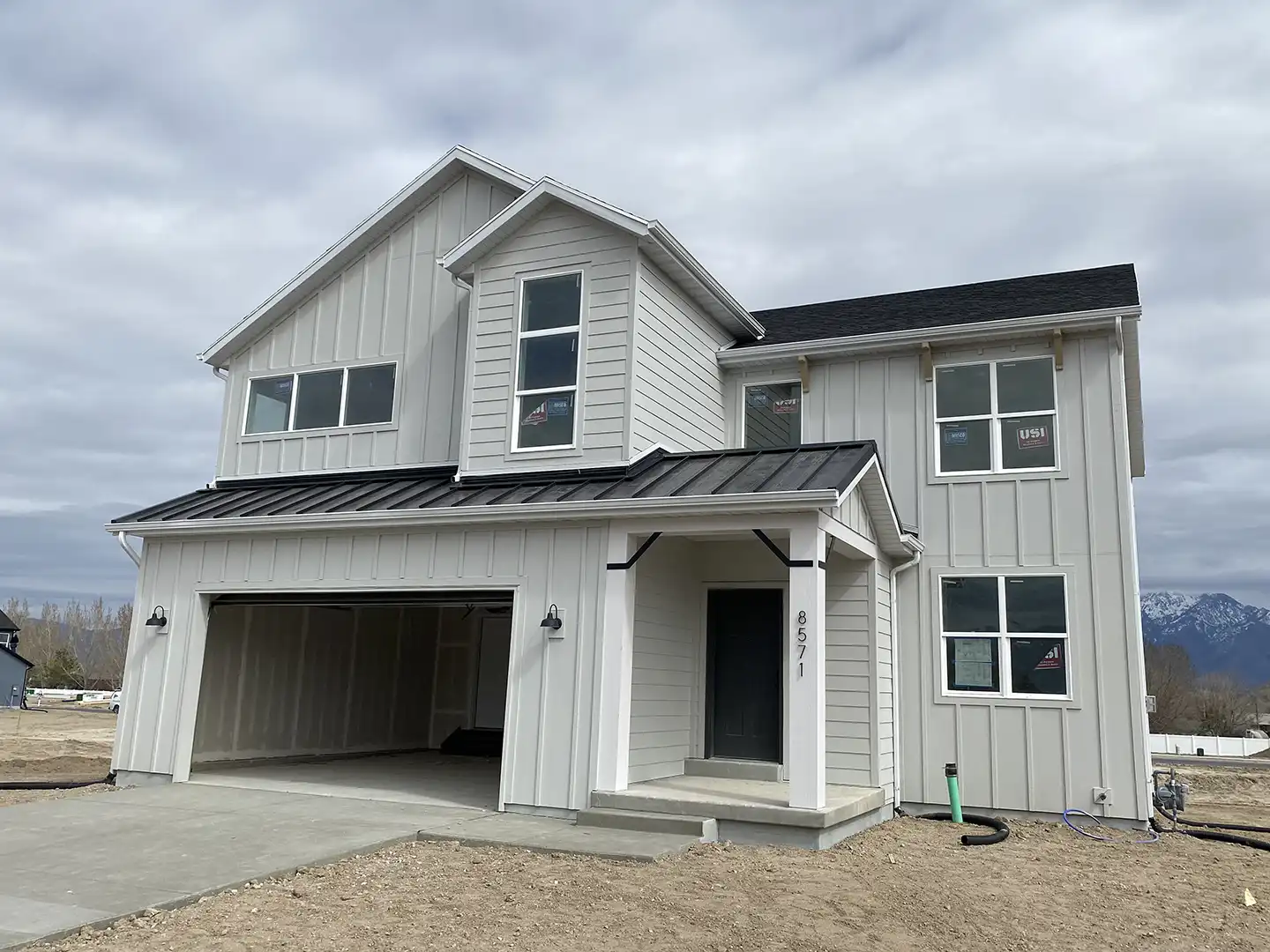 New construction modern farmhouse with light board-and-batten siding, black metal porch roof, two-car garage, address 8571, mountain view behind.