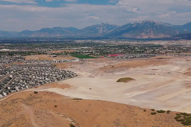 Aerial view of a graded development site with a neighborhood at left and mountains across the valley